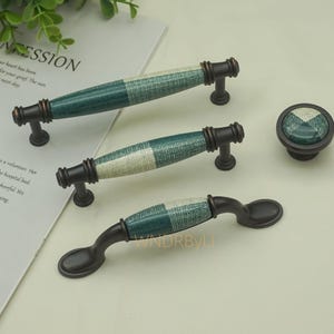 Op de afbeelding: Een set kastbeslag met donker bronzen grepen en een knop. De grepen hebben een groen en wit geblokt patroon op de keramische handvatten. De knop heeft hetzelfde patroon. Het beslag is nieuw.