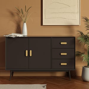 Op de afbeelding: Een donkerbruin dressoir met goudkleurige handgrepen en drie lades. De kast heeft twee deuren en staat op dunne, zwarte poten. Een witte vaas met hoog gras en een ingelijste abstracte schilderij hangen aan de muur achter de kast.