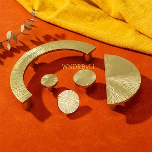 May include: A collection of gold-colored cabinet hardware, including a curved handle, a half-circle handle, and three round knobs. The hardware has a textured, linear design. The text "WNDRByLi" is visible on the orange surface.