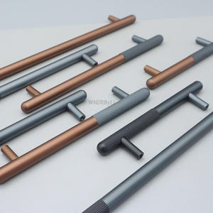 Puede incluir: Una variedad de tiradores de armario modernos en varios colores. Los tiradores tienen un diseño en forma de T con una textura acanalada en el mango. Los colores incluyen bronce, gris y negro. El hardware está dispuesto sobre una superficie gris claro.
