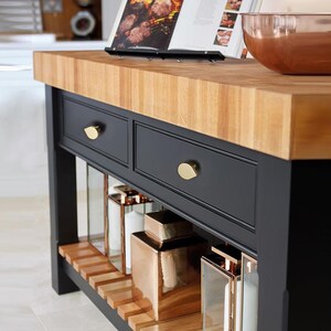 May include: A kitchen island with a wooden butcher block worktop and a black base. The island has two drawers with gold-coloured knobs, open shelving, and decorative items. A cookbook is open on the worktop, and a copper bowl is visible.