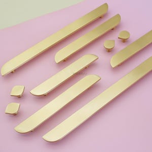 May include: A selection of gold-coloured cabinet hardware, including long bar pulls and smaller knobs, arranged on a pink surface. The hardware has a sleek, modern design with rounded edges. The image showcases various sizes and shapes of the hardware.