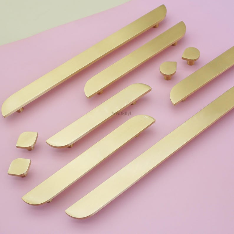 May include: A selection of gold-coloured cabinet hardware, including long bar pulls and smaller knobs, arranged on a pink surface. The hardware has a sleek, modern design with rounded edges. The image showcases various sizes and shapes of the hardware.