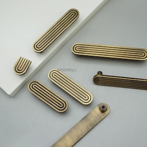 Op de afbeelding: Een verzameling antiek messing kastgrepen en knoppen. De grepen zijn rechthoekig met een groefontwerp en de knoppen hebben een U-vormig ontwerp. De hardware is gerangschikt op een wit en grijs oppervlak.