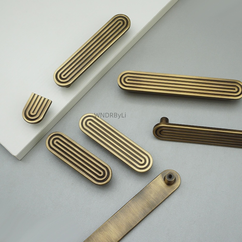 Op de afbeelding: Een verzameling antiek messing kastgrepen en knoppen. De grepen zijn rechthoekig met een groefontwerp en de knoppen hebben een U-vormig ontwerp. De hardware is gerangschikt op een wit en grijs oppervlak.