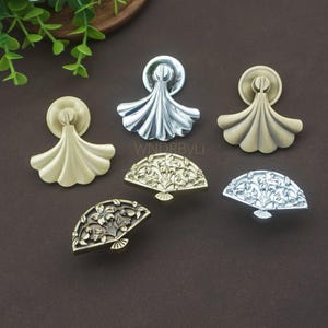 Könnte beinhalten: Eine Auswahl an dekorativen Schrankbeschlägen in verschiedenen metallischen Ausführungen. Die Auswahl umfasst fächerförmige und gewellte Griffe in Gold-, Silber- und Bronzetönen, ideal für Möbelrestaurierung oder DIY-Projekte.