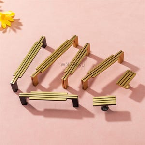 Puede incluir: Un conjunto de tiradores y pomos dorados para armarios con un dise&ntilde;o moderno. Los tiradores tienen forma rectangular con l&iacute;neas horizontales, y los pomos tienen una base circular. La ferreter&iacute;a est&aacute; sobre un fondo rosa.