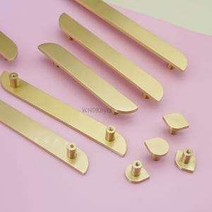 May include: Gold-coloured cabinet hardware, including long bar pulls and smaller knob pulls, are arranged on a pink surface. The bar pulls have a curved design, and the knobs have a teardrop shape. The hardware is new and unused.