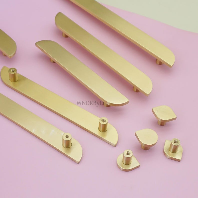 May include: Gold-coloured cabinet hardware, including long bar pulls and smaller knob pulls, are arranged on a pink surface. The bar pulls have a curved design, and the knobs have a teardrop shape. The hardware is new and unused.
