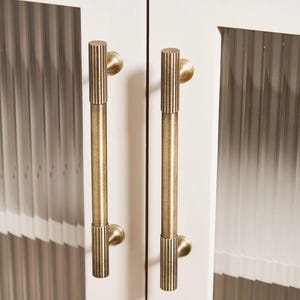 Puede incluir: Primer plano de dos tiradores de armario de lat&oacute;n antiguo. Cada tirador tiene un dise&ntilde;o vertical y acanalado en el agarre y una base circular. Los tiradores est&aacute;n montados en una puerta de armario blanca con un patr&oacute;n vertical texturizado.