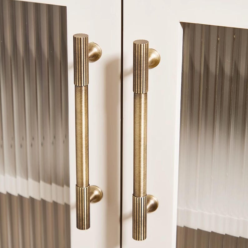 Puede incluir: Primer plano de dos tiradores de armario de lat&oacute;n antiguo. Cada tirador tiene un dise&ntilde;o vertical y acanalado en el agarre y una base circular. Los tiradores est&aacute;n montados en una puerta de armario blanca con un patr&oacute;n vertical texturizado.