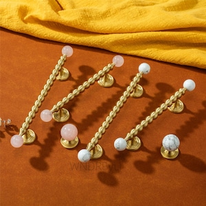 May include: A collection of brass cabinet pulls with decorative beaded stems and round, gemstone-like knobs. Some knobs are pink, others are white with gray veining. The pulls are arranged on an orange surface with a yellow fabric in the background.
