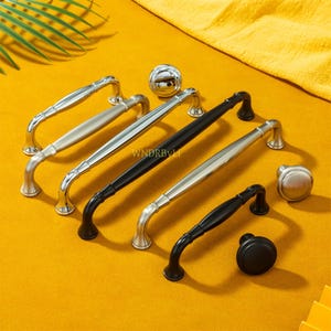 May include: An assortment of cabinet hardware, including handles and knobs, in silver and black finishes. The handles have a curved design, and the knobs are round. The hardware is displayed on a yellow surface.