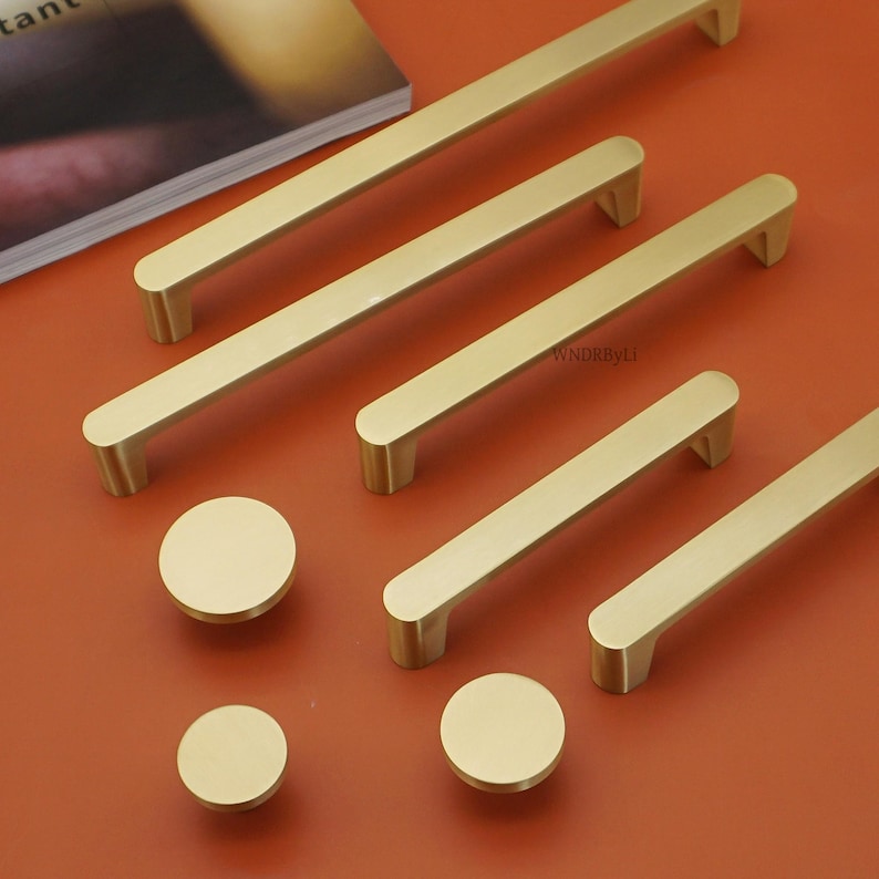 May include: Gold-toned cabinet hardware set, including several long bar handles and round knobs. The handles are rectangular with rounded edges and are arranged on an orange surface. The knobs are circular and have a matching gold finish. This set is ideal for updating kitchen or bathroom cabinets.