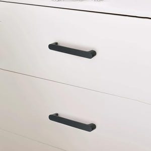 May include: Close-up of a white dresser with two black, rectangular drawer pulls. The pulls are horizontally mounted and have a rounded design. The dresser has a clean, modern aesthetic.