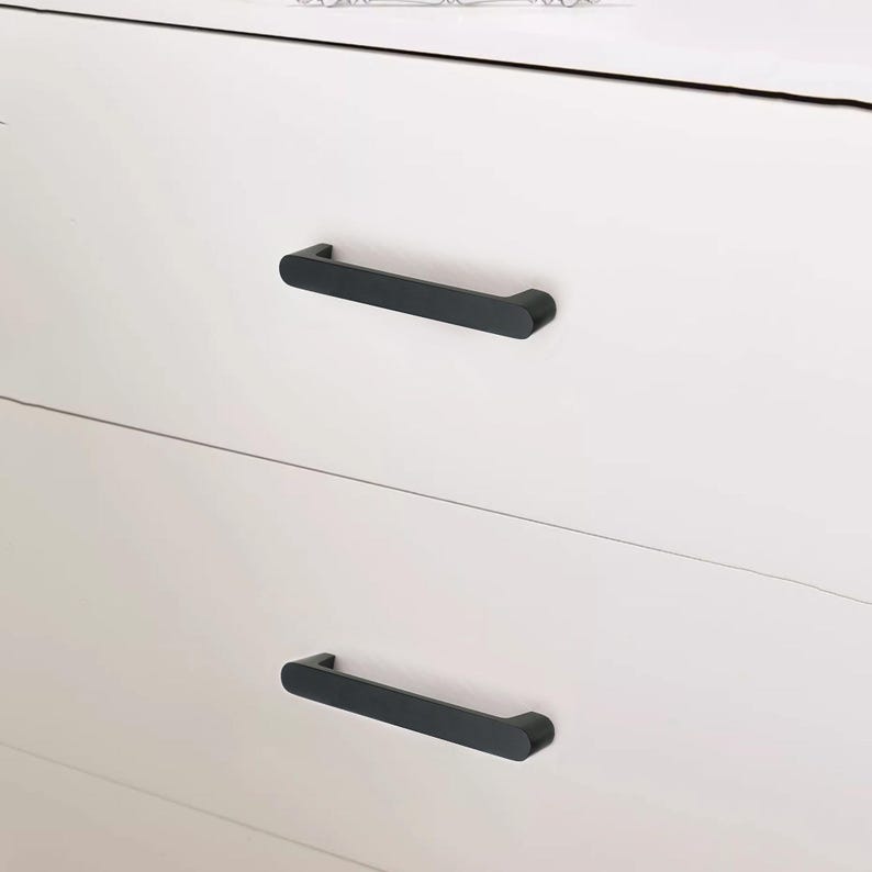 May include: Close-up of a white dresser with two black, rectangular drawer pulls. The pulls are horizontally mounted and have a rounded design. The dresser has a clean, modern aesthetic.