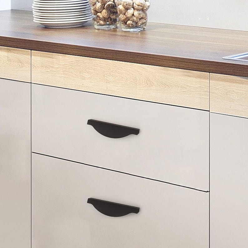 May include: Two black cabinet pulls are mounted on a white cabinet drawer. The pulls have a curved, minimalist design. The cabinet is part of a kitchen setup, with a wooden countertop and other kitchen items visible in the background.