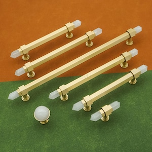 May include: A collection of gold-toned cabinet pulls and a knob with clear crystal accents. The pulls are rectangular with crystal ends, and the knob is a round crystal shape. The hardware is displayed on a green and orange background.