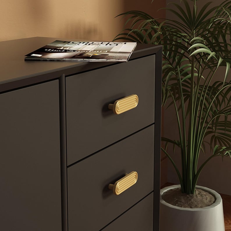 Op de afbeelding: Donkergrijze ladekast met drie lades, elk met een goudkleurige, rechthoekige handgreep. Een tijdschrift ligt bovenop. Een potplant is rechts van de lades te zien.