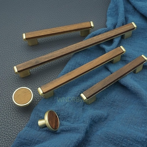 Peut inclure: Ensemble de quincaillerie de meubles en bois, comprenant des poignées et des boutons. Le matériel présente un design rectangulaire en bois de noyer avec des accents de couleur laiton. Les boutons sont ronds avec une base en laiton.