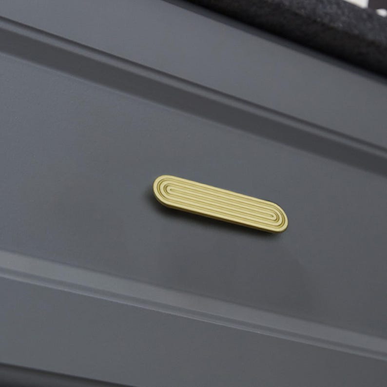 Op de afbeelding: Een goudkleurige, ovaalvormige kastgreep met een groefontwerp. De greep is gemonteerd op een donkergrijze kastdeur. De hardware heeft een strakke, moderne esthetiek, geschikt voor diverse meubelstijlen.