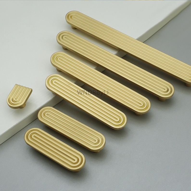 Op de afbeelding: Een verzameling goudkleurige meubelbeslag, waaronder verschillende langwerpige handgrepen en een enkele knop. Het beslag heeft een gegroefd, lineair ontwerp binnen een afgeronde rechthoekige vorm. De stukken zijn gerangschikt op een lichtgrijze en witte ondergrond.