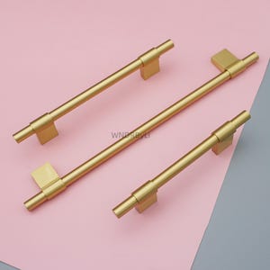 Op de afbeelding: Drie goudkleurige kastgrepen met een rechthoekige basis en een cilindrische handgreep. De grepen zijn geplaatst op een roze en grijze achtergrond. De tekst "WNDRBYLI" is zichtbaar op de roze achtergrond.