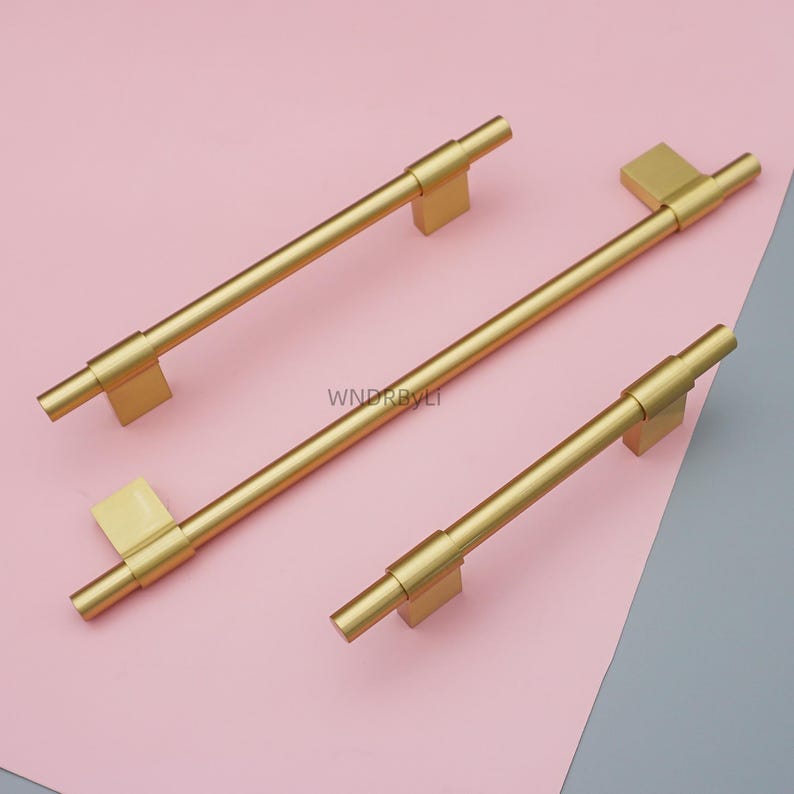 Op de afbeelding: Drie goudkleurige kastgrepen met een rechthoekige basis en een cilindrische handgreep. De grepen zijn geplaatst op een roze en grijze achtergrond. De tekst "WNDRBYLI" is zichtbaar op de roze achtergrond.