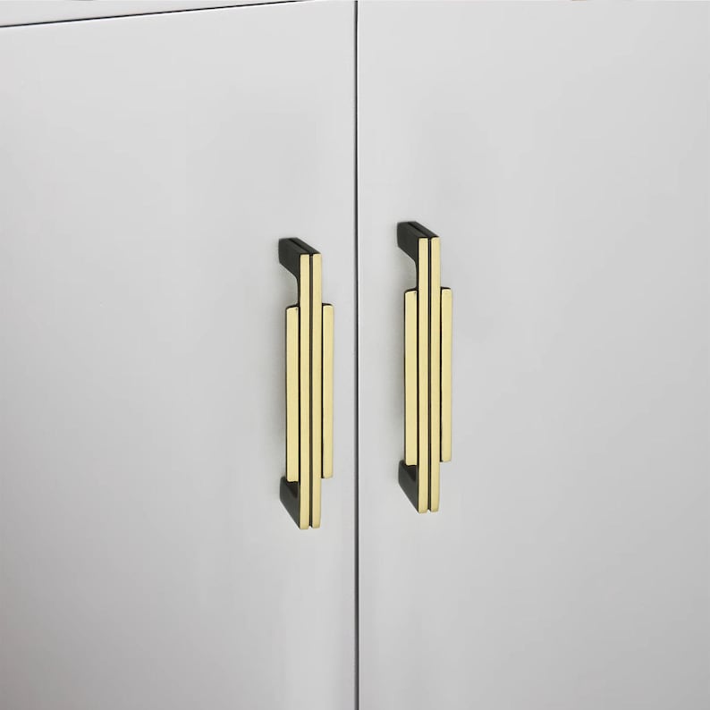 Puede incluir: Dos tiradores de armario con parte superior e inferior negras y un dise&ntilde;o vertical dorado. Los tiradores est&aacute;n montados en una puerta de armario blanca. El dise&ntilde;o es geom&eacute;trico y moderno.