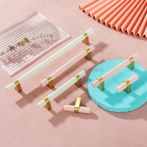 May include: A collection of cabinet pulls in various sizes and colors. The pulls have a cylindrical design with gold-colored hardware and are available in white, pink, and mint green. The pulls are displayed on a pink and blue surface.