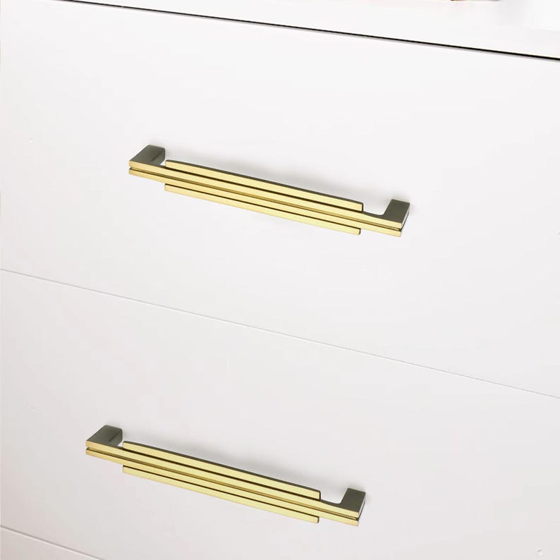 Puede incluir: Primer plano de una c&oacute;moda blanca con dos cajones, cada uno con un tirador rectangular dorado. Los tiradores tienen un dise&ntilde;o en capas, a&ntilde;adiendo un toque moderno a los muebles. Los cajones son limpios y minimalistas.