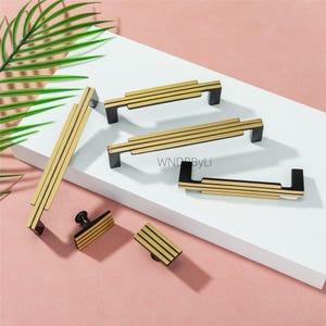 Puede incluir: Una colecci&oacute;n de herrajes dorados y negros para gabinetes, que incluyen manijas y perillas. El herraje presenta un dise&ntilde;o lineal con rayas horizontales doradas sobre una base negra. La imagen est&aacute; sobre un fondo blanco y rosa.