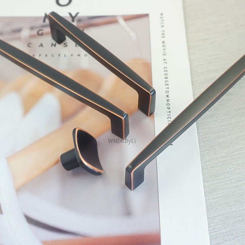 May include: Close-up of several black cabinet pulls and a knob with copper-coloured accents. The hardware is displayed on a magazine page. The pulls are rectangular with a slightly curved design. The knob is square with rounded edges.