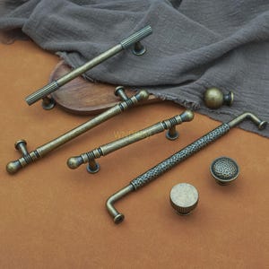 May include: An assortment of antique brass cabinet hardware, including handles and knobs. The handles vary in design, with ribbed, hammered, and smooth textures. The hardware is displayed on a brown surface with a gray cloth in the background.