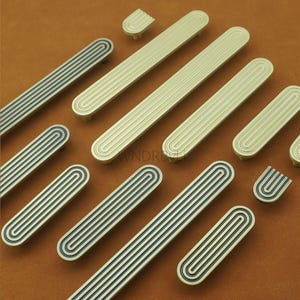 May include: An assortment of gold and black cabinet pulls and knobs. The pulls are long and oval-shaped with a series of vertical grooves. Some pulls have black stripes. The knobs are small and rectangular. The hardware is arranged on a brown surface.