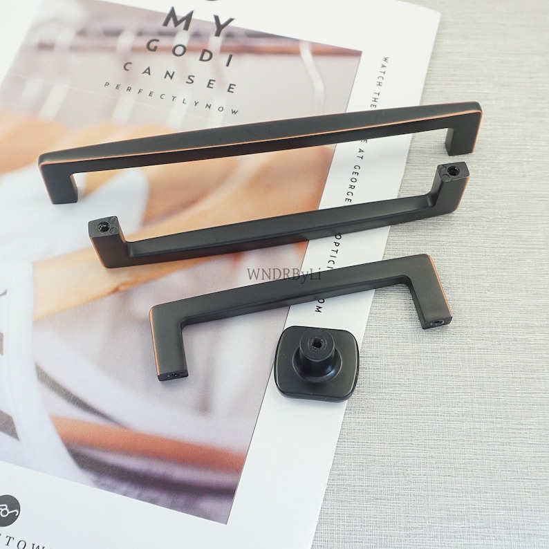 May include: A selection of black cabinet handles and a knob with copper-coloured accents. The handles are rectangular with a bar design. The knob is square with a circular centre. The items are on a white paper with text.