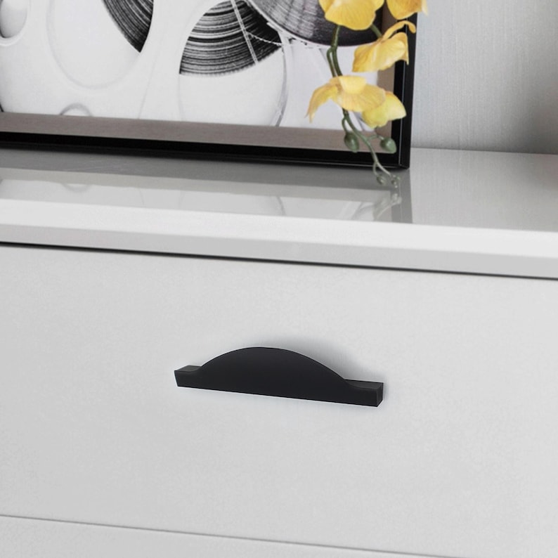 May include: A close-up of a white dresser drawer with a black, curved handle. The drawer is part of a white dresser. A framed black and white picture and yellow flowers are in the background.