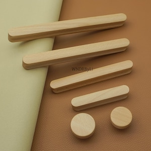 May include: A collection of wooden cabinet hardware, including rectangular pulls and round knobs. The hardware is made of light-colored wood and is arranged on a tan and cream-colored surface. The text "WNDRByLi" is visible.