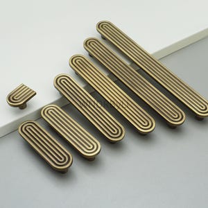 Op de afbeelding: Een verzameling antieke messing kastgrepen en knoppen. De grepen zijn rechthoekig met parallelle groeven, terwijl de knop een gebogen ontwerp heeft. De hardware is gerangschikt op een licht oppervlak, wat hun vintage esthetiek laat zien.