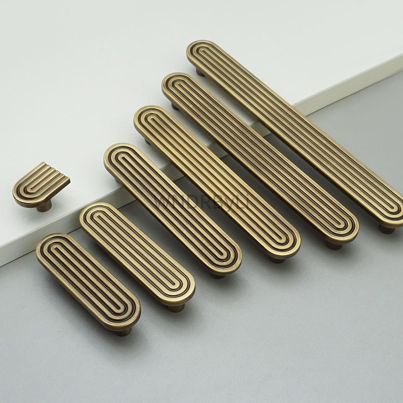 Op de afbeelding: Een verzameling antieke messing kastgrepen en knoppen. De grepen zijn rechthoekig met parallelle groeven, terwijl de knop een gebogen ontwerp heeft. De hardware is gerangschikt op een licht oppervlak, wat hun vintage esthetiek laat zien.