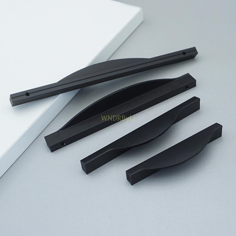 May include: Four black cabinet pulls in varying sizes. Each pull has a curved, ergonomic design with a matte finish. The pulls are made of metal and are shown on a light gray surface. The text "WNDRB" is visible on one of the pulls.
