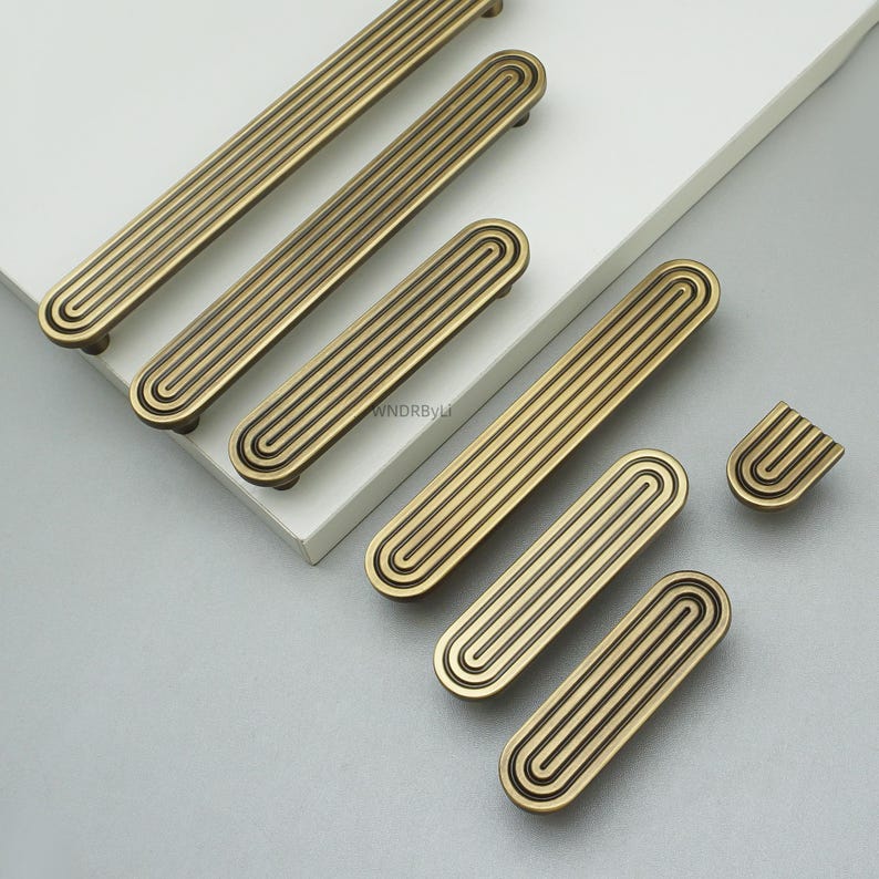 Op de afbeelding: Een verzameling antiek messing kastgrepen en -knoppen. De hardware heeft een gegroefd, rechthoekig ontwerp met afgeronde uiteinden. De grepen en knoppen zijn in verschillende maten, met een warme, gouden afwerking. De hardware wordt gepresenteerd op een wit oppervlak.