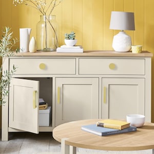 May include: Cream-colored sideboard with two drawers and two doors, featuring gold-toned hardware. The open door reveals storage space. A round wooden coffee table sits in front. Decorative items include a lamp, vase, and small potted plant.