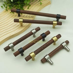 Peut inclure: Plusieurs poignées de meubles en bois avec des détails métalliques. Les poignées présentent des barres en bois brun foncé avec des garnitures métalliques argentées, noires et dorées. Elles sont présentées sur une surface claire, mettant en valeur leur design et leur finition.