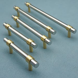 Op de afbeelding: Een set van zes kastgrepen met een modern design. Elke greep heeft een geborstelde zilveren staaf met goudkleurige accenten aan de uiteinden en de basis. De grepen zijn diagonaal geplaatst tegen een lichtblauwe achtergrond, wat hun strakke, eigentijdse stijl laat zien.