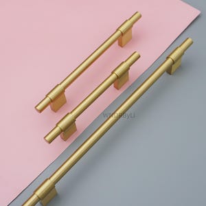 Op de afbeelding: Vier goudkleurige kastgrepen, elk met een rechthoekige basis en een cilindrische handgreep, tegen een roze en grijze achtergrond. De handgrepen hebben een dikker middengedeelte. De grepen zijn van metaal en hebben een matte afwerking. Ongeveer 15 cm lang.