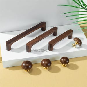 May include: A collection of wooden cabinet hardware, including several dark brown rectangular handles and round knobs. The knobs have a gold-colored base. The handles vary in size, and the knobs are uniform in size.