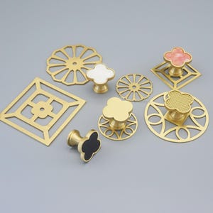 May include: A selection of decorative cabinet knobs and pulls in various shapes. The knobs have gold-toned metal bases with clover, flower, and geometric designs. Some have white, pink, or black accents. The hardware is displayed on a grey background.