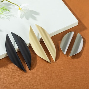May include: A collection of leaf-shaped cabinet pulls in various finishes: matte black, brushed gold, and brushed silver. The hardware is arranged on a white surface with a brown leather-like background, showcasing their design and texture.