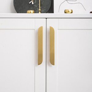 May include: Close-up of a white cabinet with two gold-coloured, vertical handles. The handles have a rounded top and are mounted on the doors. The cabinet is part of a larger piece of furniture, with decorative items on top.